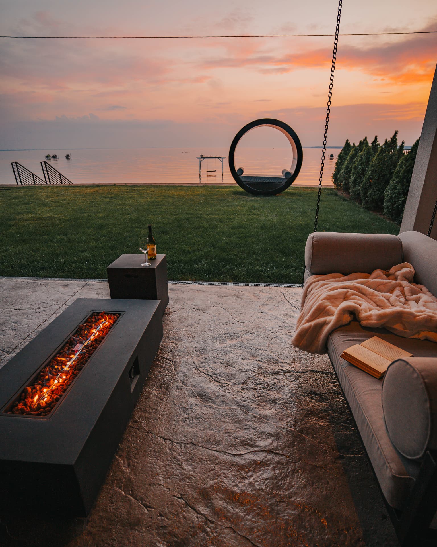 Covered patio with linear fire table and hanging daybed facing the lakefront lawn