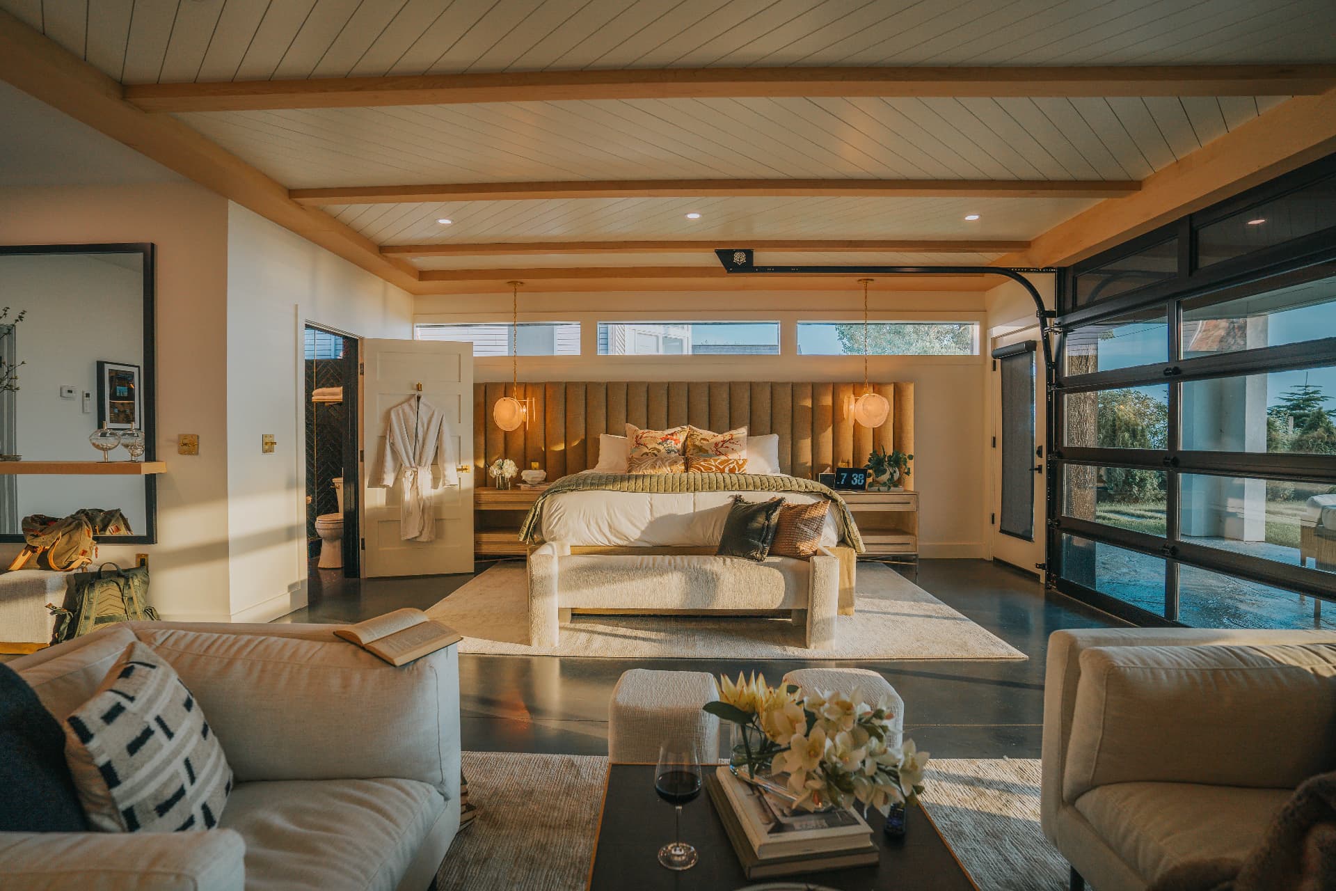First-floor primary suite with channel-tufted headboard, exposed beams, and glass garage-style door to the lawn